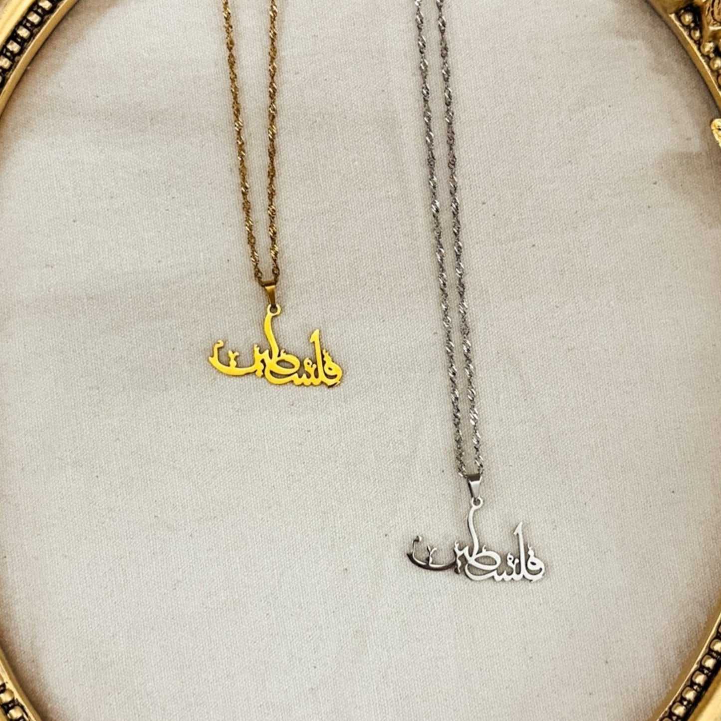 Gold and silver Falasteen necklaces in Arabic script displayed in an ornate gold butterfly frame. The Palestine necklaces, symbolizing solidarity, are set against a soft neutral fabric backdrop, surrounded by white flowers, candles, and crystal beads, showcasing their elegant design and craftsmanship. Ideal for those supporting Palestine through meaningful jewelry.