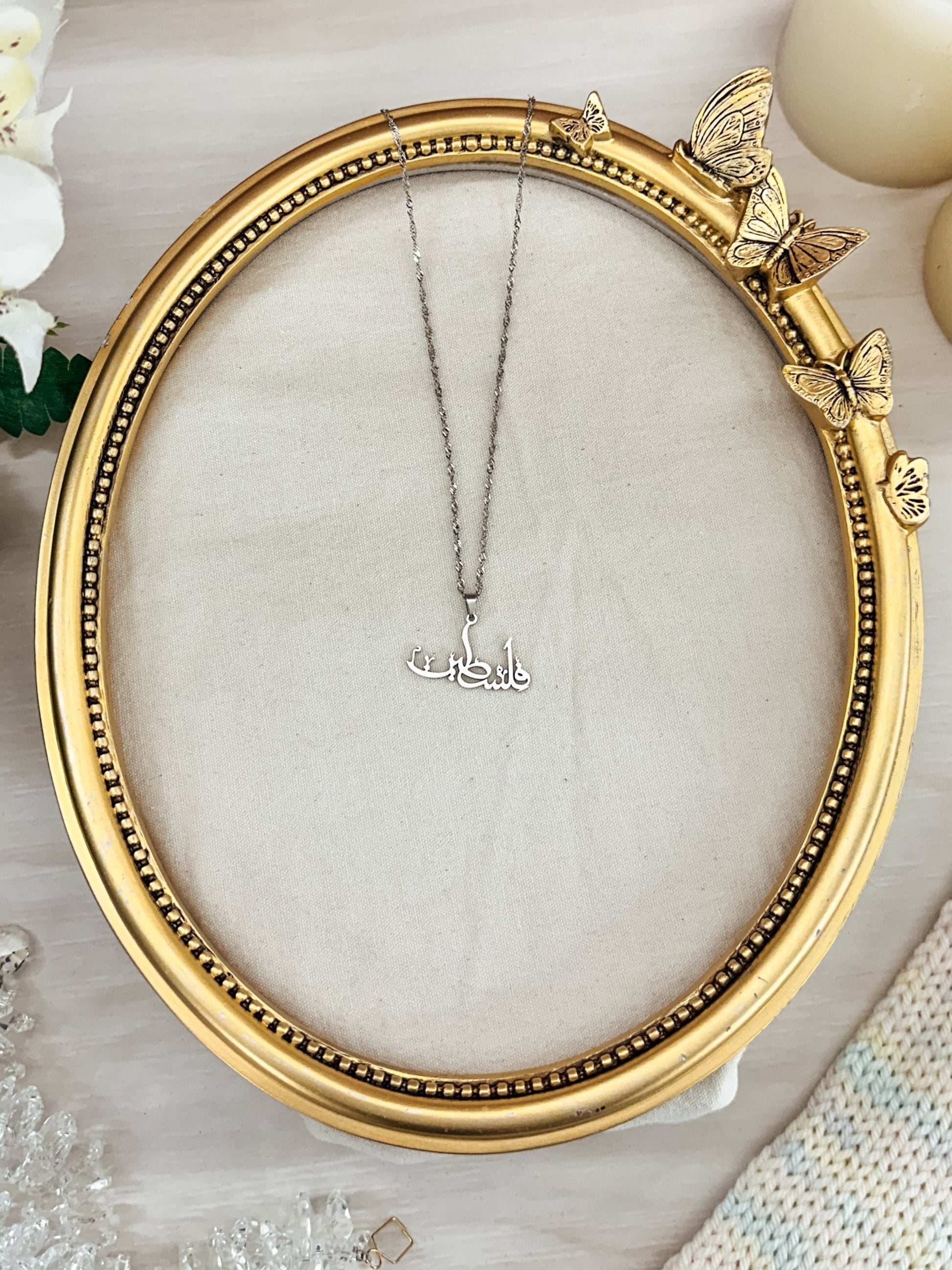 Silver Falasteen necklace in Arabic script displayed in a gold ornate butterfly frame. The Arabic Palestine necklace is set against a soft neutral fabric, surrounded by white flowers, candles, and crystal beads, highlighting its elegant design and craftsmanship. Perfect for showing solidarity and support for Palestine.