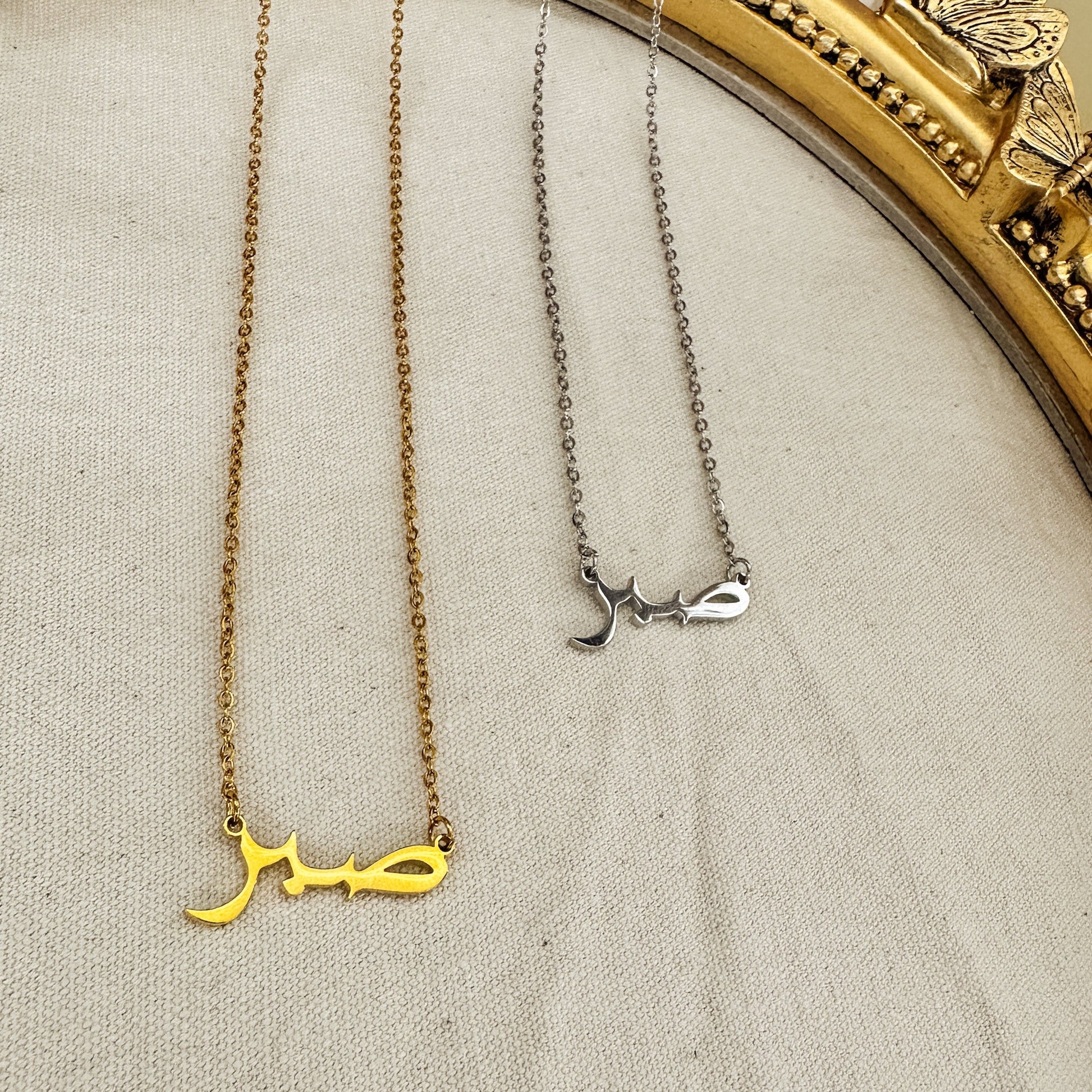 Gold and silver Sabr necklaces displayed in Arabic script, meaning “Patience,” set against a neutral fabric backdrop within an ornate gold butterfly frame. These elegant, hypoallergenic, and waterproof necklaces symbolize strength and resilience, perfect for everyday wear. Ideal for those who value patience and seek meaningful jewelry.