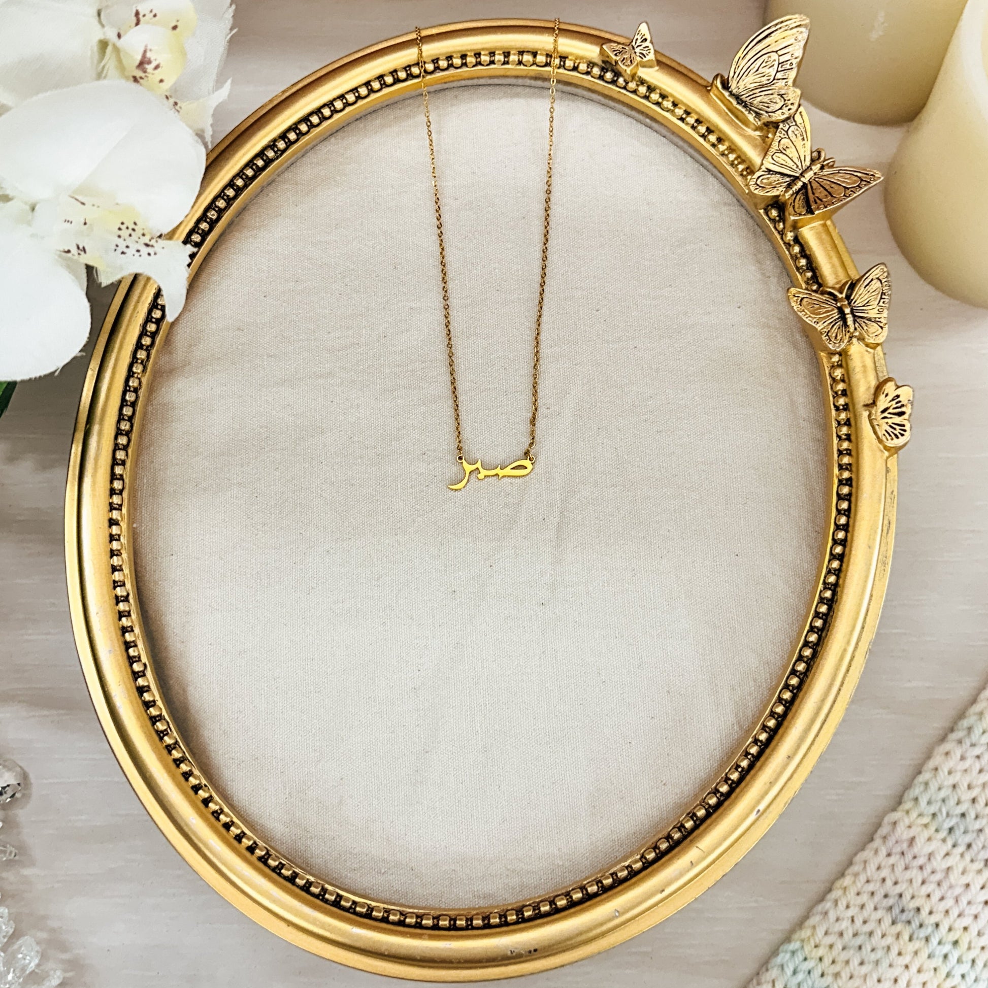 gold Sabr necklaces displayed in Arabic script, meaning “Patience,” set against a neutral fabric backdrop within an ornate gold butterfly frame. These elegant, hypoallergenic, and waterproof necklaces symbolize strength and resilience, perfect for everyday wear. Ideal for those who value patience and seek meaningful jewelry.