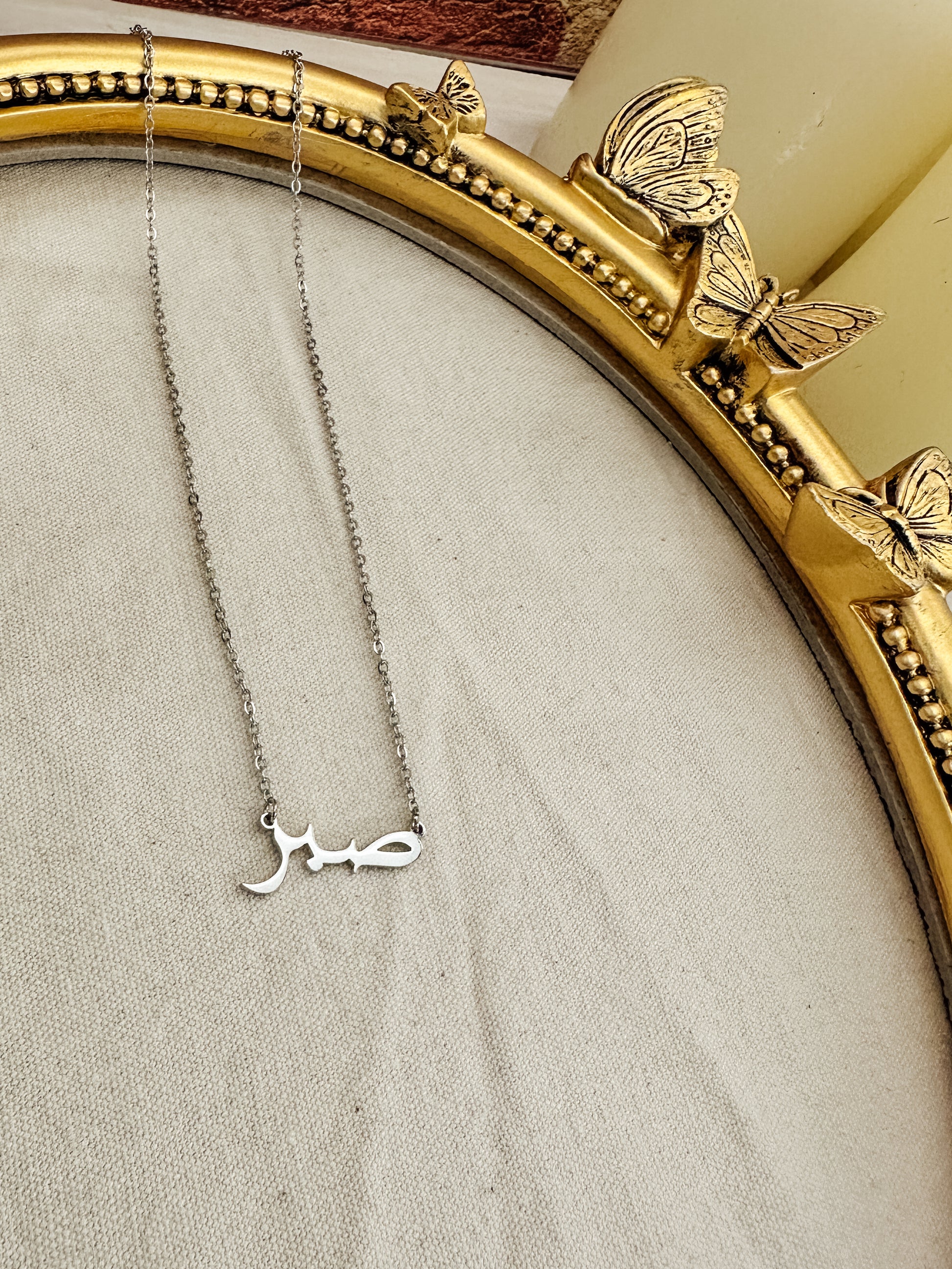 silver Sabr necklaces displayed in Arabic script, meaning “Patience,” set against a neutral fabric backdrop within an ornate gold butterfly frame. These elegant, hypoallergenic, and waterproof necklaces symbolize strength and resilience, perfect for everyday wear. Ideal for those who value patience and seek meaningful jewelry.